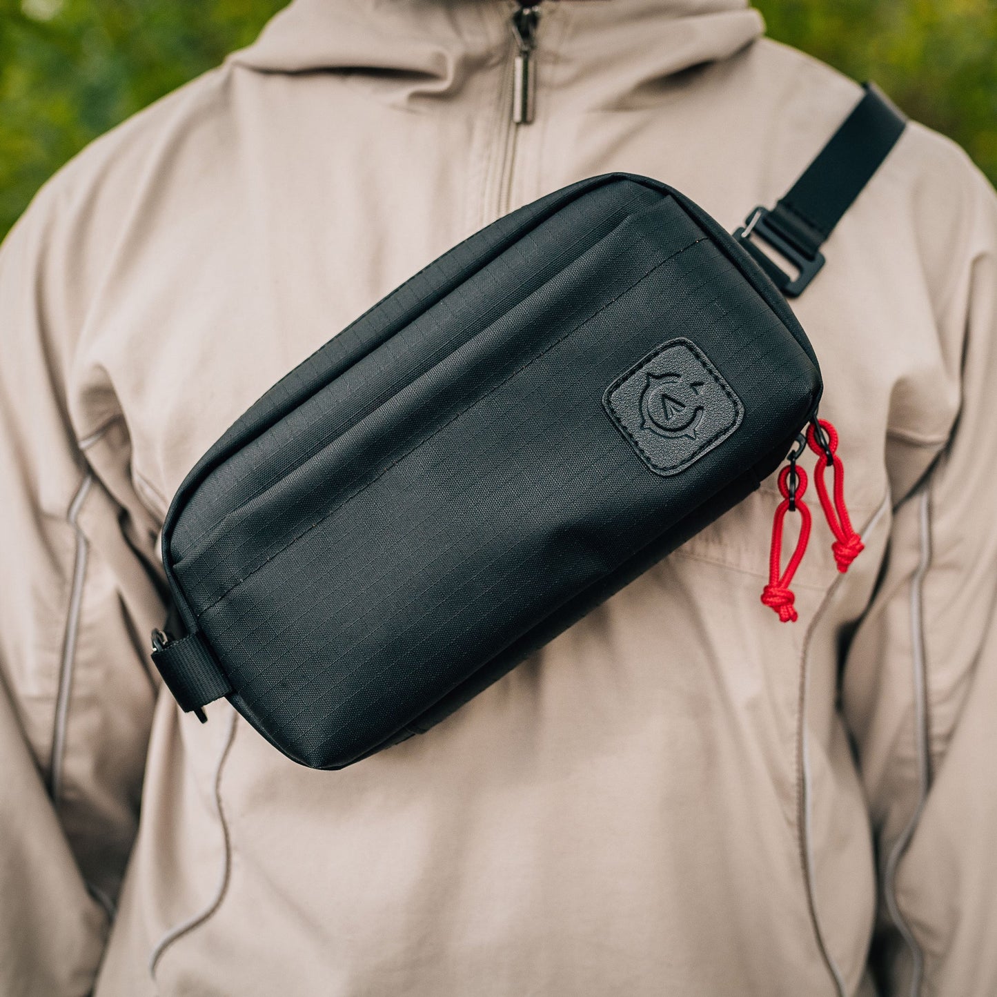 A person wearing a beige jacket with a Cancha Sling Bag, featuring red zipper pull tabs, weather-resistant pockets, and the Cancha logo on the front.