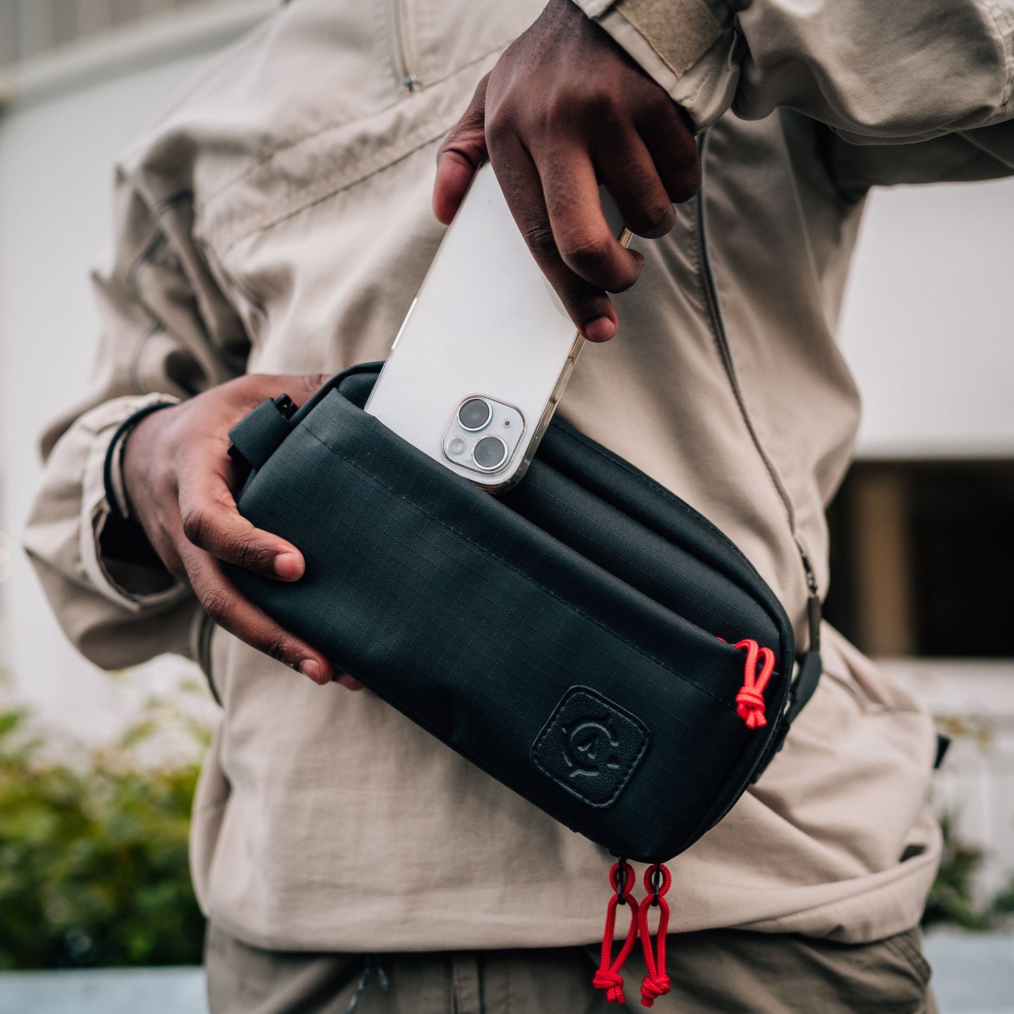 A person in a beige jacket puts a smartphone into a black Cancha Sling Bag with red zipper pulls, demonstrating its adaptable design.