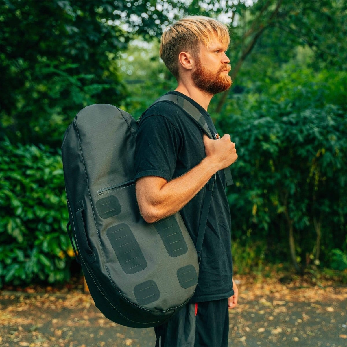 A person with a beard and short hair carries Cancha's Original Racquet Bag (25L) on one shoulder, a large black backpack made from lightweight water-repellent materials, while standing outdoors near greenery.