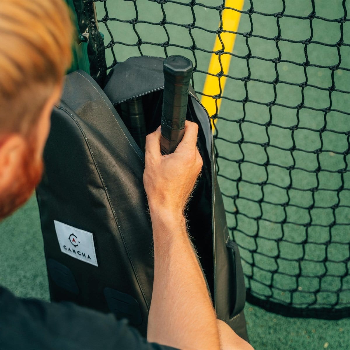 A person with light brown hair is placing a tennis racket into a sleek, black Original Racquet Bag (25L) by Cancha next to a netted fence on a tennis court. The bag, made from lightweight water-repellent materials, offers customizable add-ons for all your gear needs.