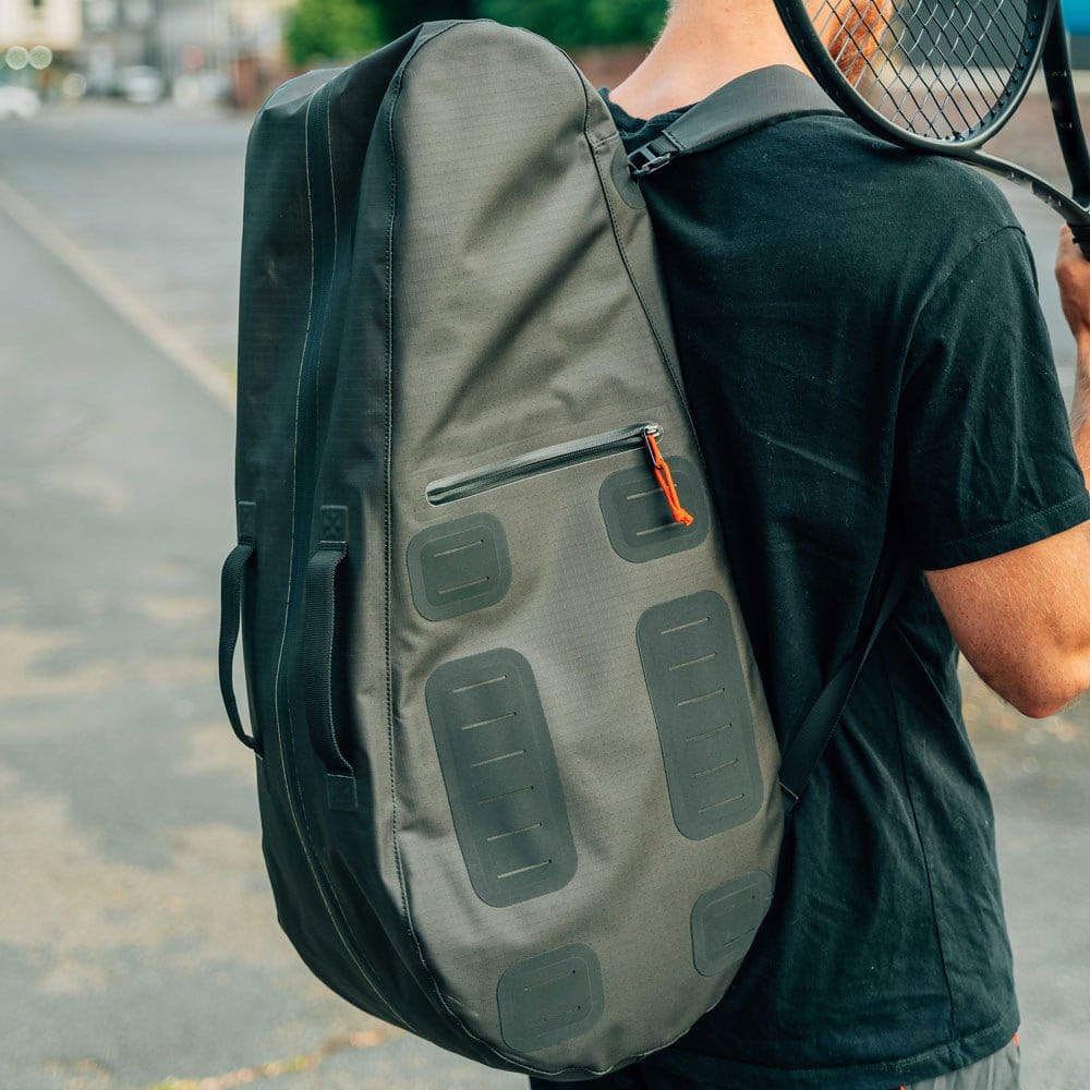 A person wearing a black t-shirt carries the Cancha Original Racquet Bag (25L), a large, lightweight, water-repellent tennis racquet bag, on their back and holds a tennis racket over their shoulder while standing on a paved area.