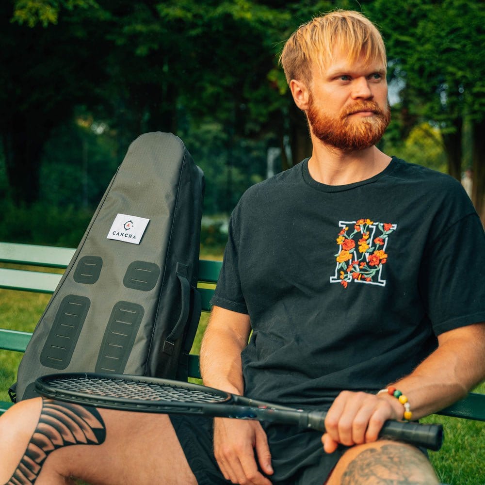 A man with a beard sits on a bench holding a tennis racket, with an Original Racquet Bag (25L) by Cancha beside him.