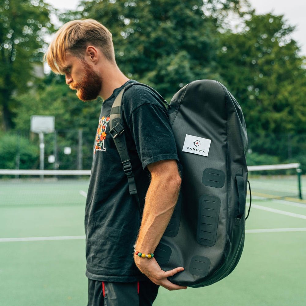 A bearded man sporting a black t-shirt is seen carrying the Cancha Original Racquet Bag (25L) over his shoulder. Crafted from lightweight, water-repellent materials, the large black bag stands out as he stands on an outdoor tennis court with a net and trees in the background.