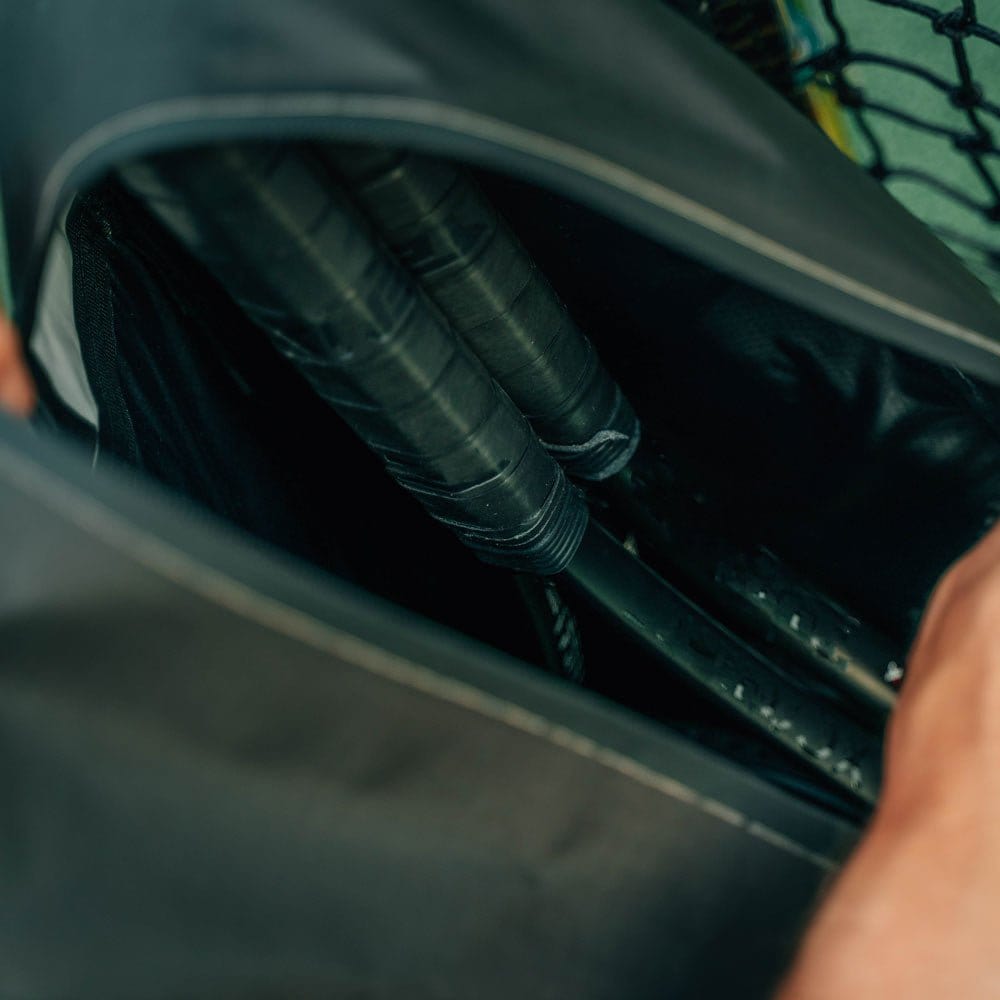 An unzipped Cancha Original Racquet Bag (25L) in black reveals two black racket handles inside, showcasing its lightweight, water-repellent materials.