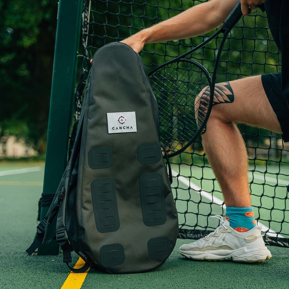 Person on a tennis court holding a lightweight, water-repellent 25L Original Racquet Bag by Cancha, with a net in the background.