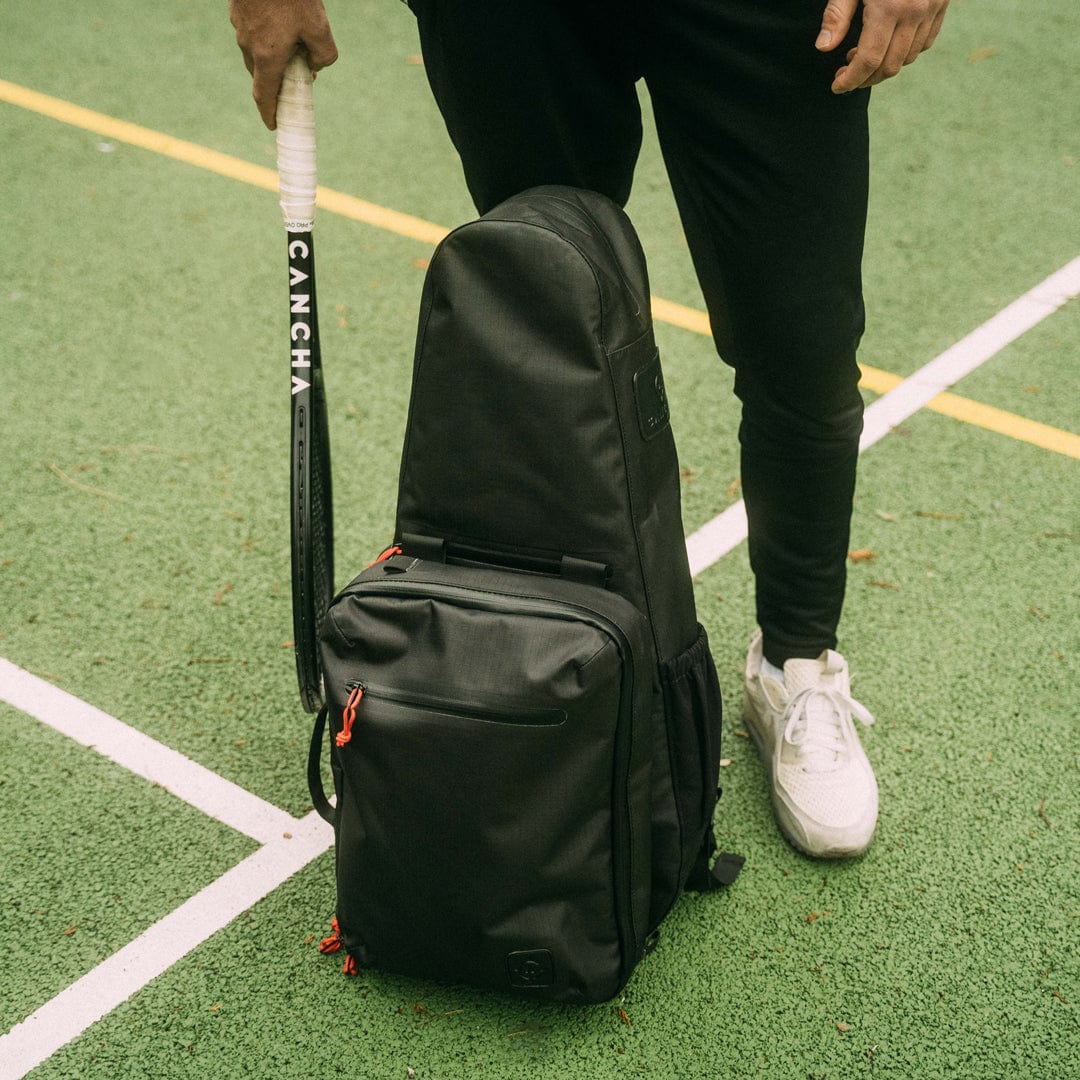 A person standing on a tennis court holding a tennis racket in their left hand, with a Cancha Racquet Bag Voyager made of weatherproof materials placed on the ground.