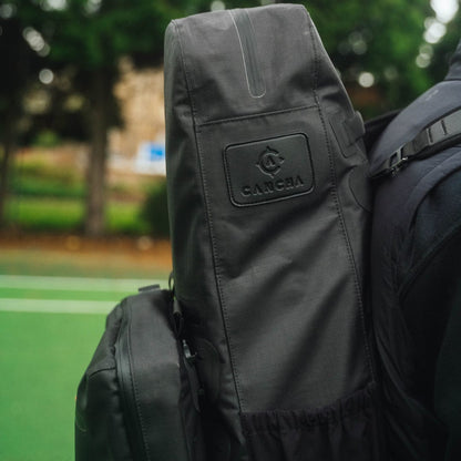 A Cancha Racquet Bag Voyager in dark gray is seen carried on a person's back. The background shows a blurred outdoor setting with green trees and a lawn, highlighting its weatherproof materials perfect for tennis travel.