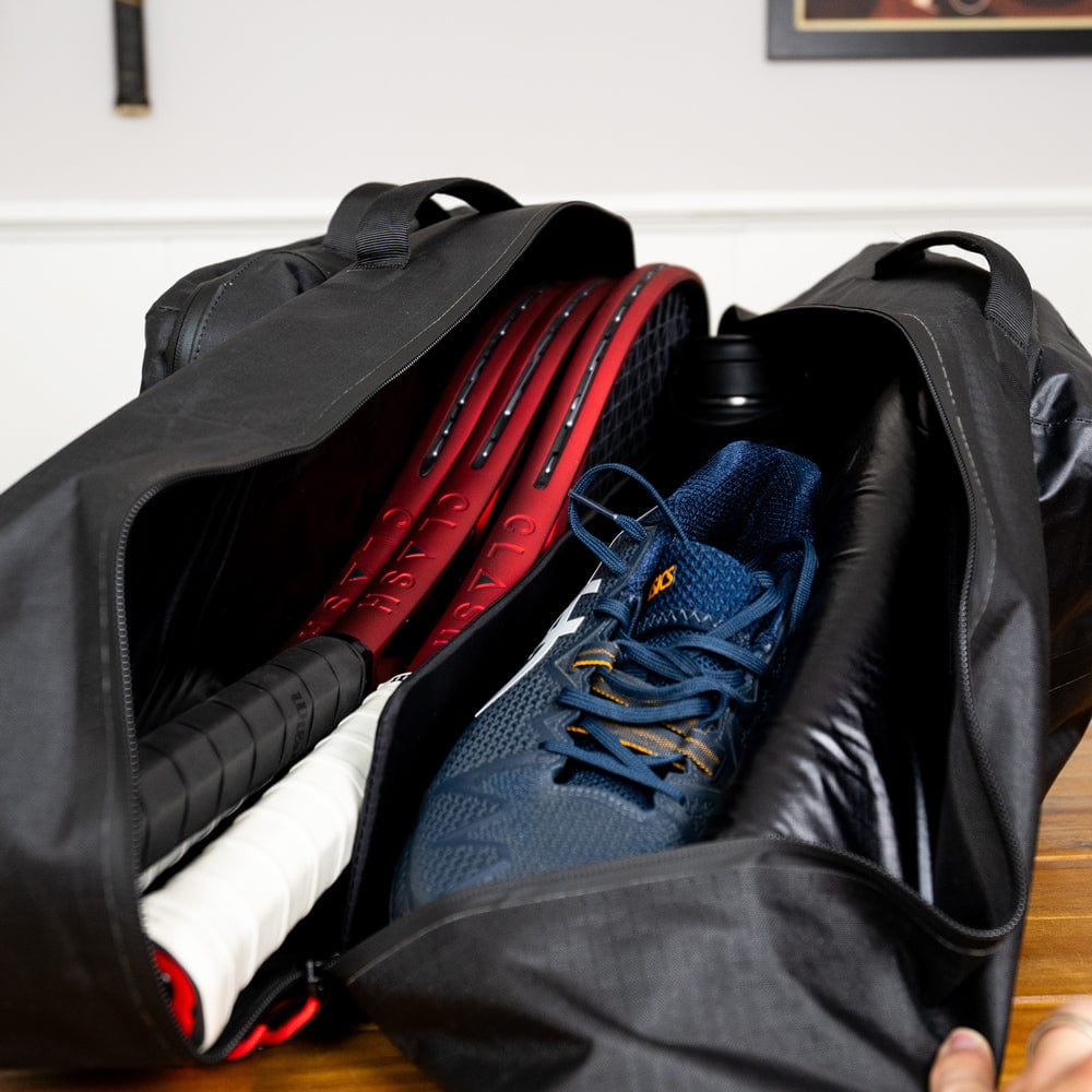 The **Original Racquet Bag (25L)** by **Cancha** sits on a wooden surface, partially open to reveal two tennis rackets with red frames, a pair of blue sports shoes, and a black water bottle inside. This black sports bag is crafted from lightweight, water-repellent materials for ultimate convenience and durability.