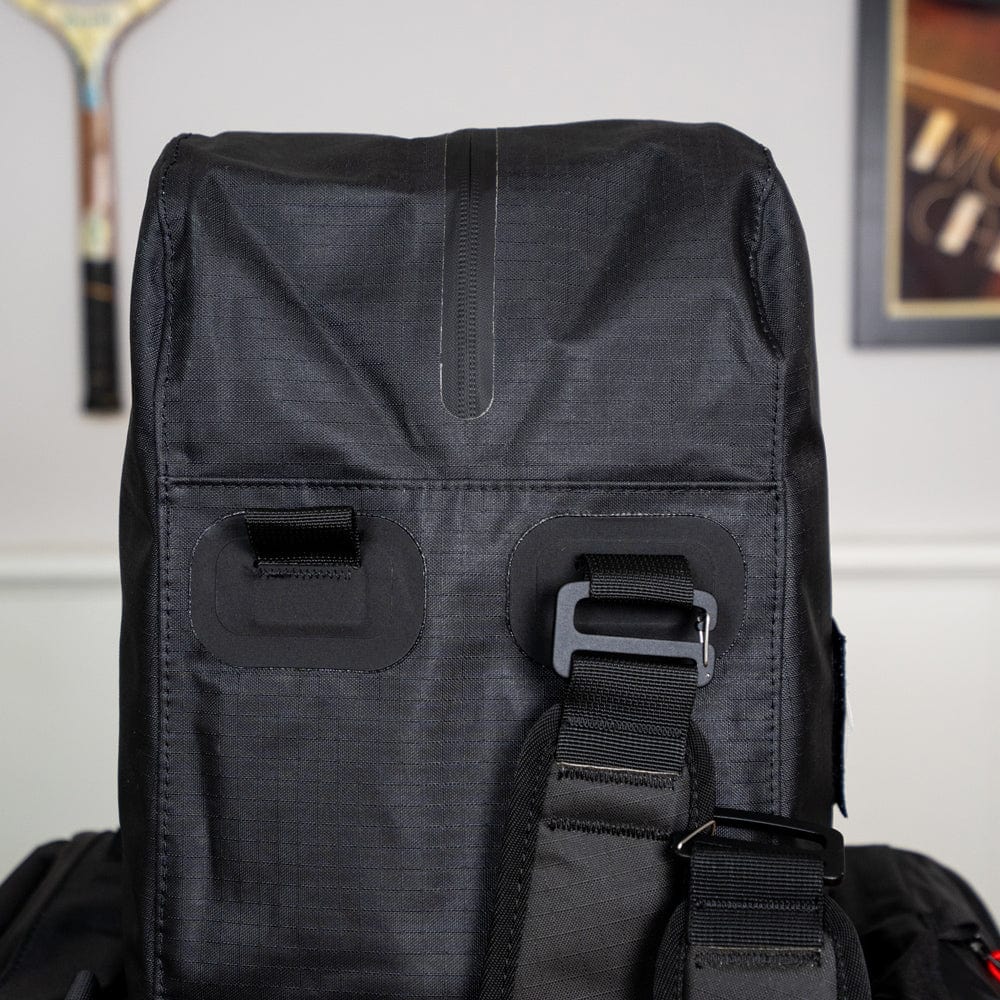 Front view of the black Original Racquet Bag (25L) by Cancha, featuring a strap and buckles, made from lightweight water-repellent materials, set against a white wall with a framed picture in the background.