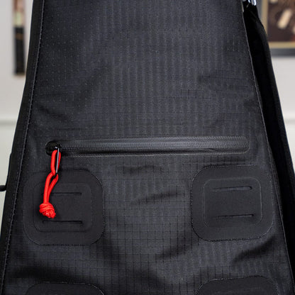 Close-up of the Cancha Original Racquet Bag (25L) in black, showcasing a zippered pocket with a red zipper pull and a grid-like fabric texture. Crafted from lightweight, water-repellent materials, it offers customizable add-ons for ultimate convenience.