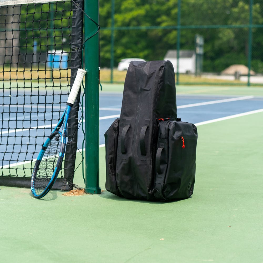 A Cancha Original Racquet Bag (25L) made from lightweight, water-repellent materials and a blue tennis racket leaning against a net post on an outdoor tennis court.