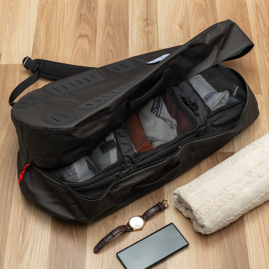 A black Cancha Original Racquet Bag (25L) is partially unzipped, revealing packed clothes. Next to it are a rolled white towel, a smartphone, and a wristwatch on the wooden floor. The bag features customizable add-ons, ensuring you have everything you need for your journey.