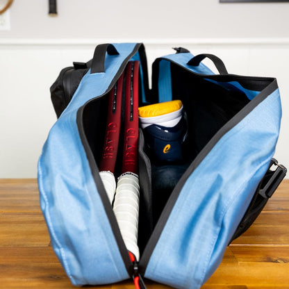 A Cancha Racquet Bag Voyager, in blue, lies open on a wooden surface, revealing two red tennis racquets, a pair of sneakers with orange accents, and other gear made from weatherproof materials.