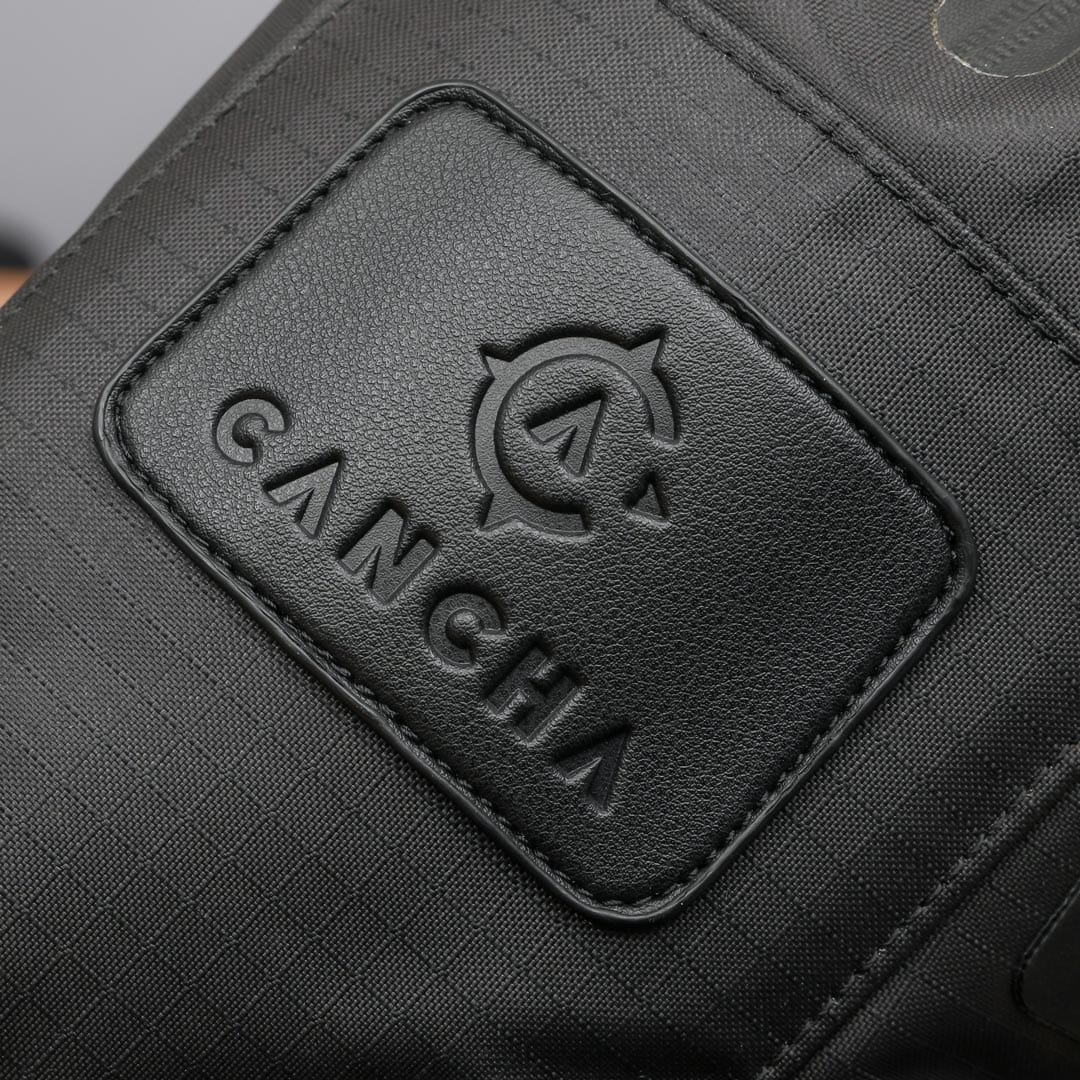 Close-up of the Racquet Bag Voyager from Cancha, featuring a stitched leather patch with the word "CANCHA" and a compass logo. Crafted from weatherproof materials, this black fabric bag is perfect for tennis travel.