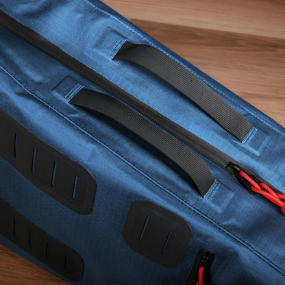 Close-up image of the Cancha Racquet Bag Voyager, featuring a blue weatherproof design with black handles and red zipper pulls, placed on a wooden surface.