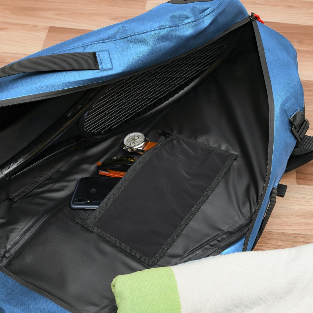 The Cancha Racquet Bag Voyager, with its zipper open, reveals a partially visible tennis racket, a wristwatch, a pair of glasses, a smartphone, and a piece of green and white fabric on a wooden floor. Perfect for tennis travel, it’s made from weatherproof materials to protect your gear.