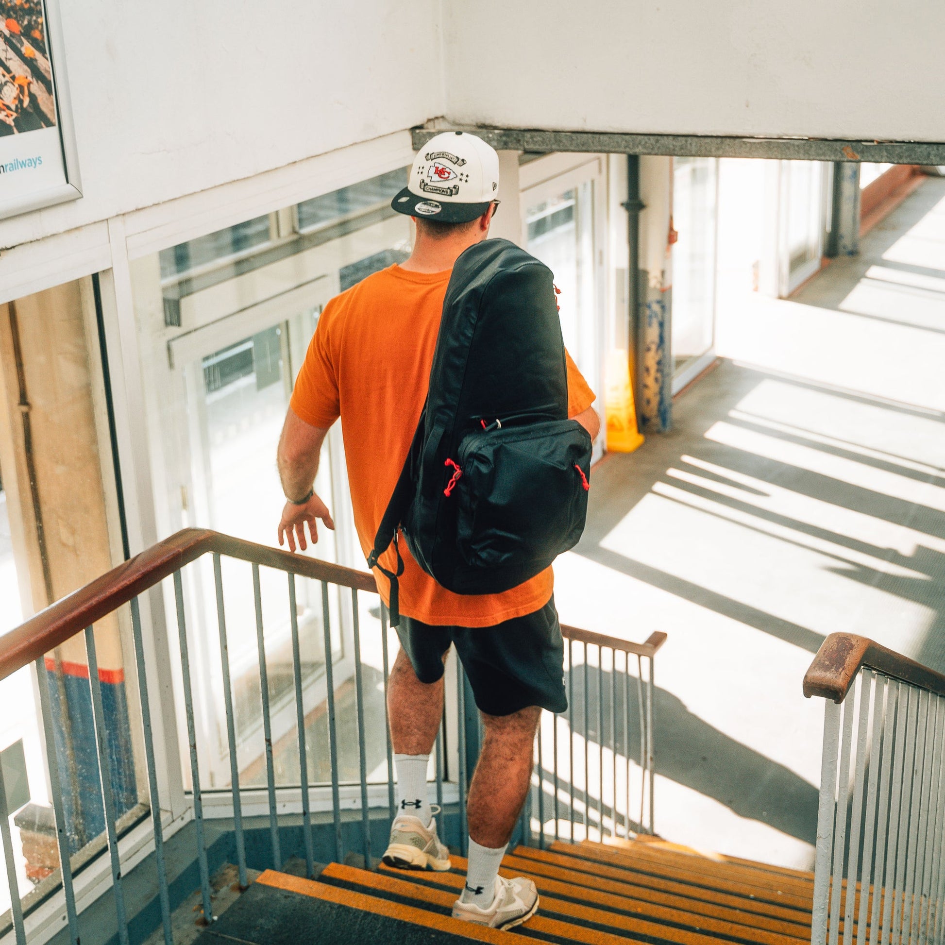 A person in an orange shirt, black shorts, and a white cap walks down the stairs with the Cancha Racquet Bag Voyager. Sunlight streams through windows, casting shadows on the stairs as they prepare for tennis travel.