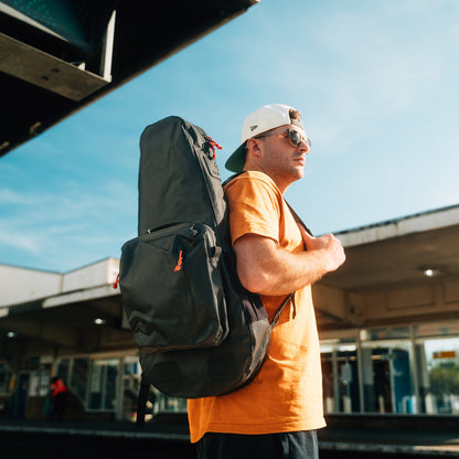 A man wearing a white cap, orange t-shirt, and sunglasses stands at a train station carrying the Cancha Racquet Bag Voyager with modular add-ons.