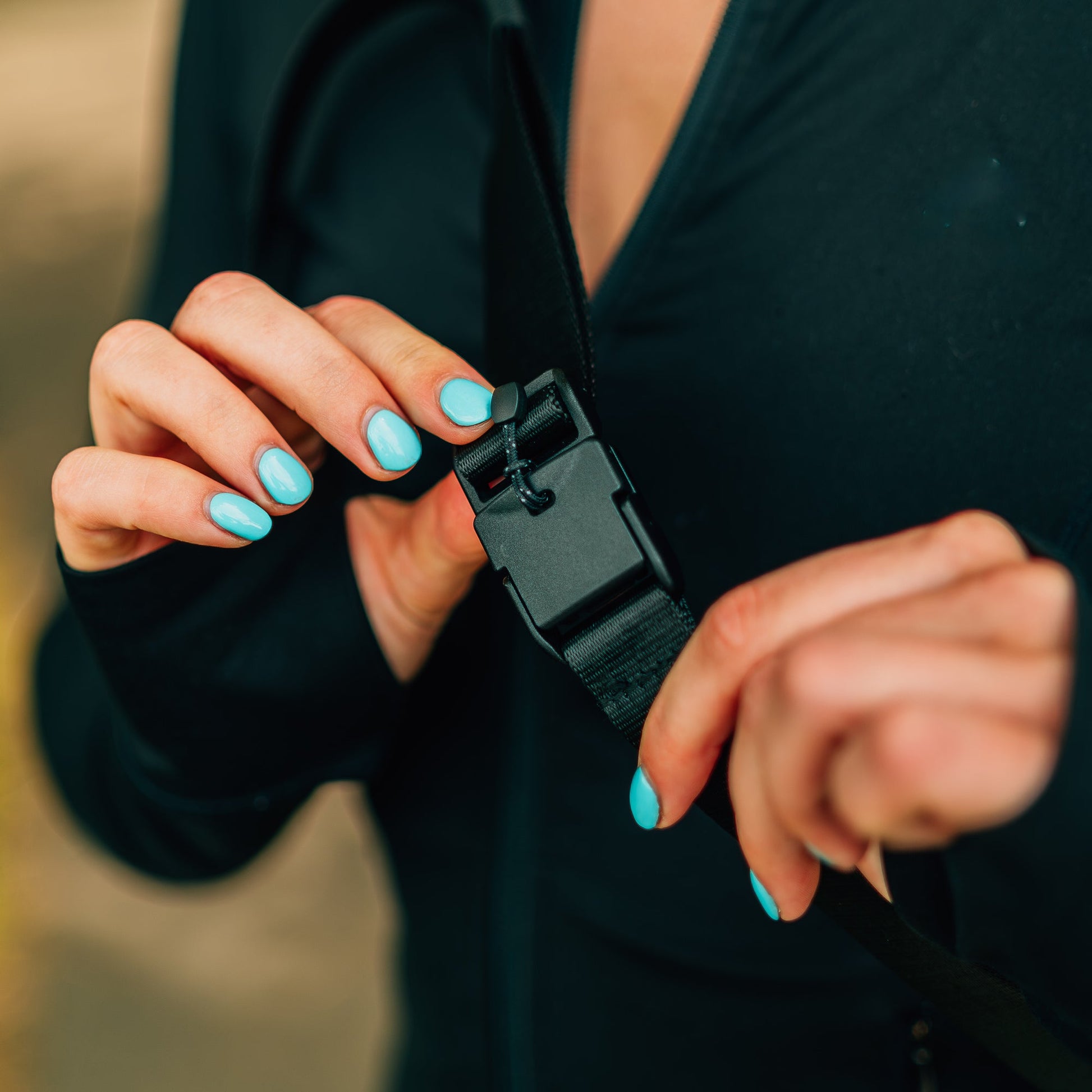 Close-up of a person with blue fingernail polish fastening a black buckle on a strap of the Cancha Sling Bag with an adaptable design, while wearing a black long-sleeve top.