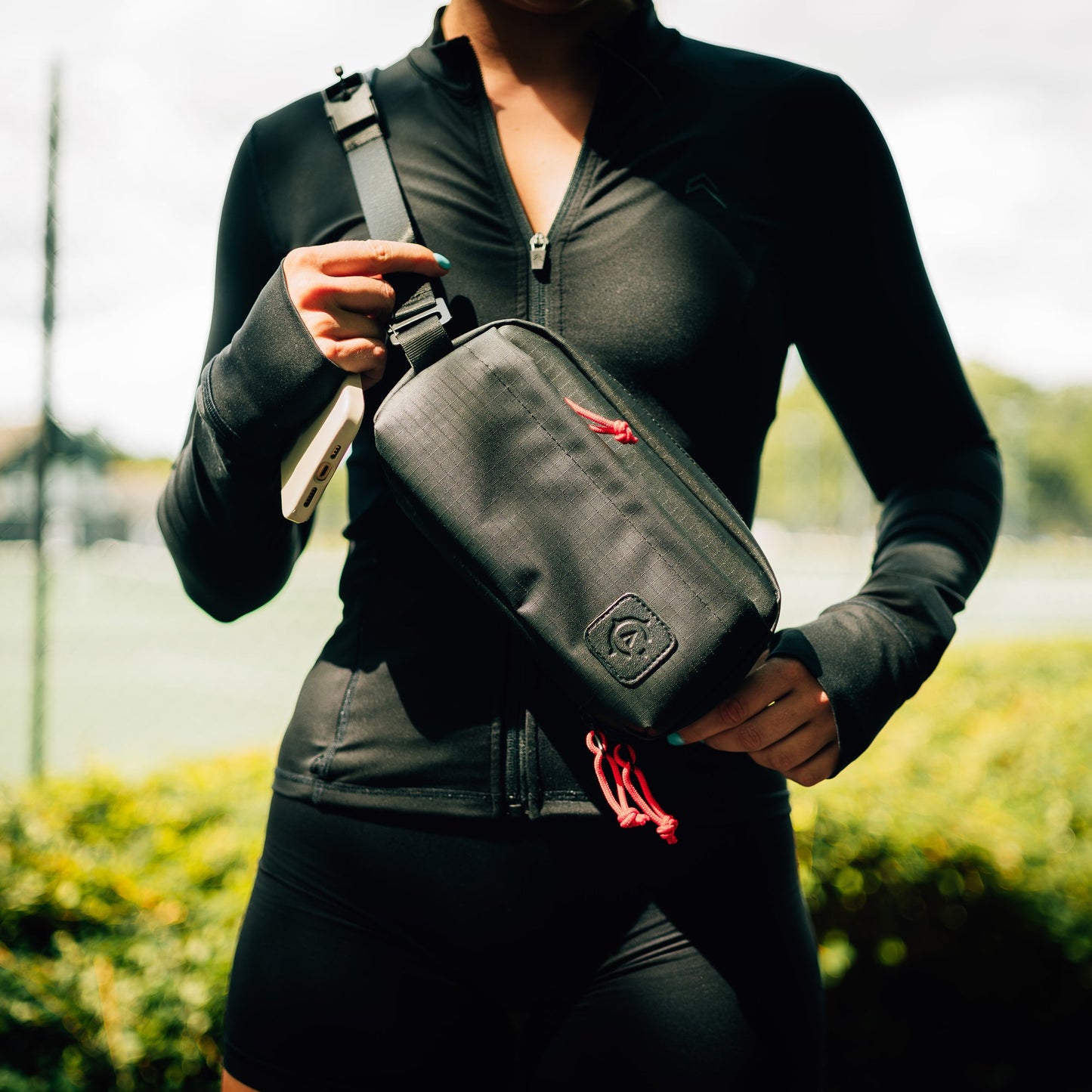 A person in a black athletic outfit is holding a Cancha Sling Bag with red zipper pulls, showcasing its travel functionality, in front of a blurred outdoor background.