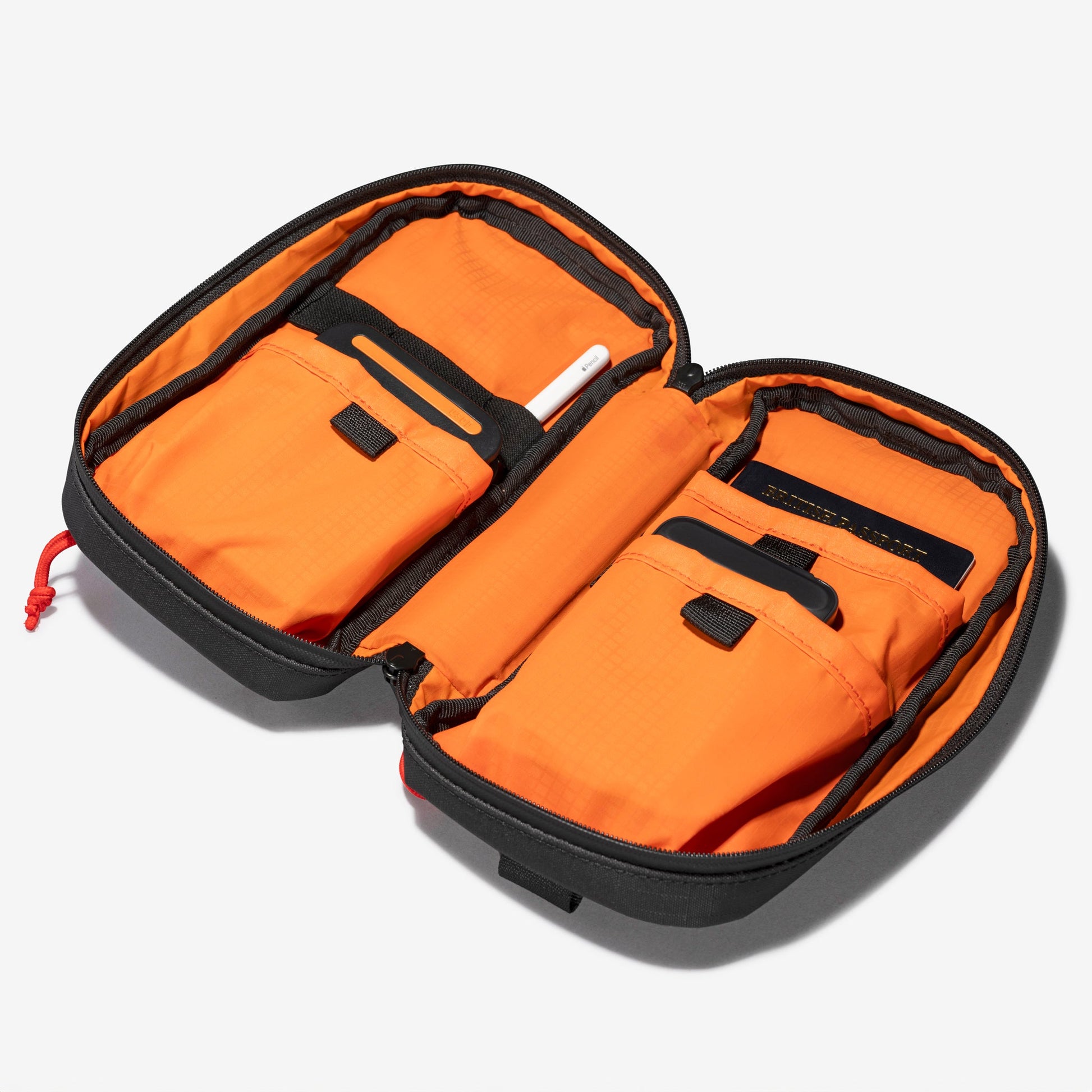 The Cancha Sling Bag in black and orange, opened to reveal two main compartments with weather-resistant pockets, holding a white pen and a small black notebook. Its adaptable design makes it perfect for various needs.